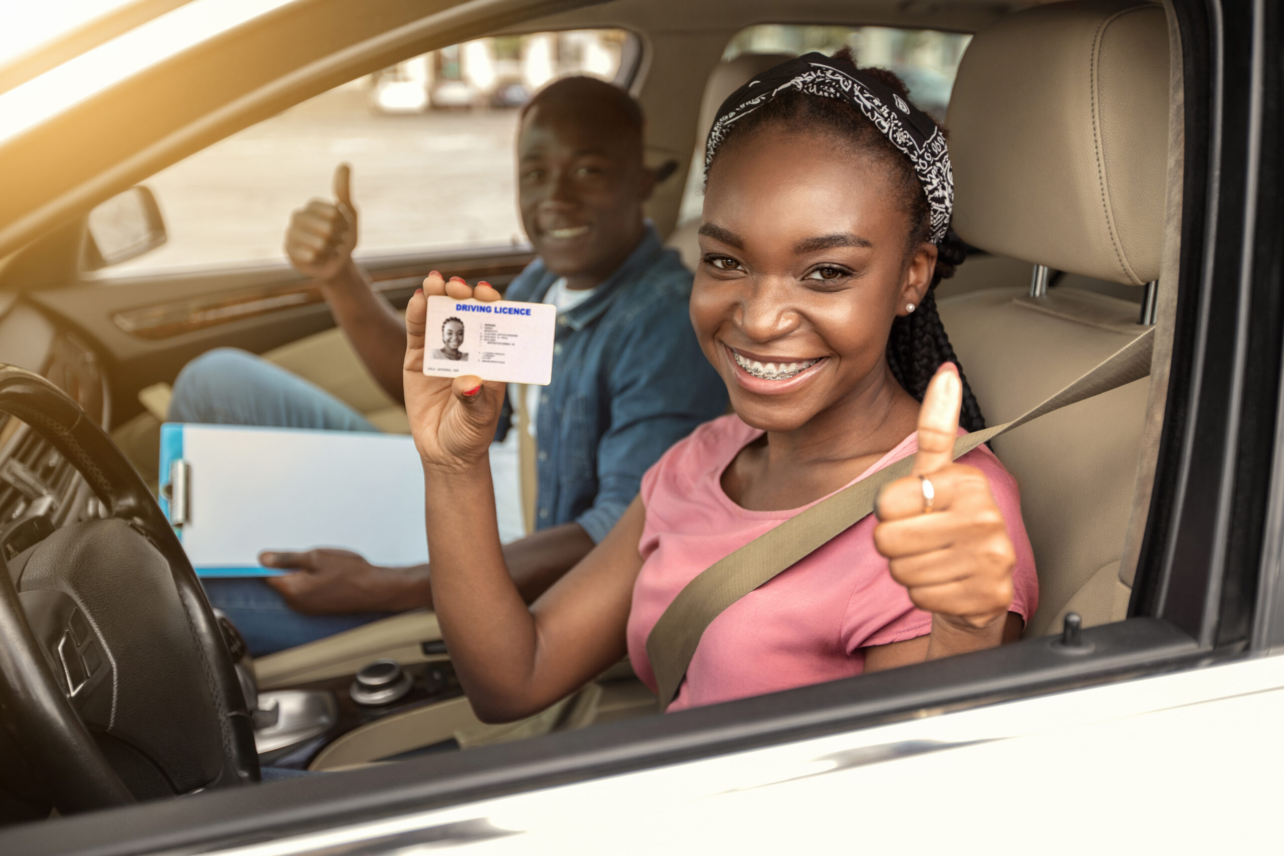 Happy african girl with driving license and her instructor showing thumb up inside car, woman passed driving test