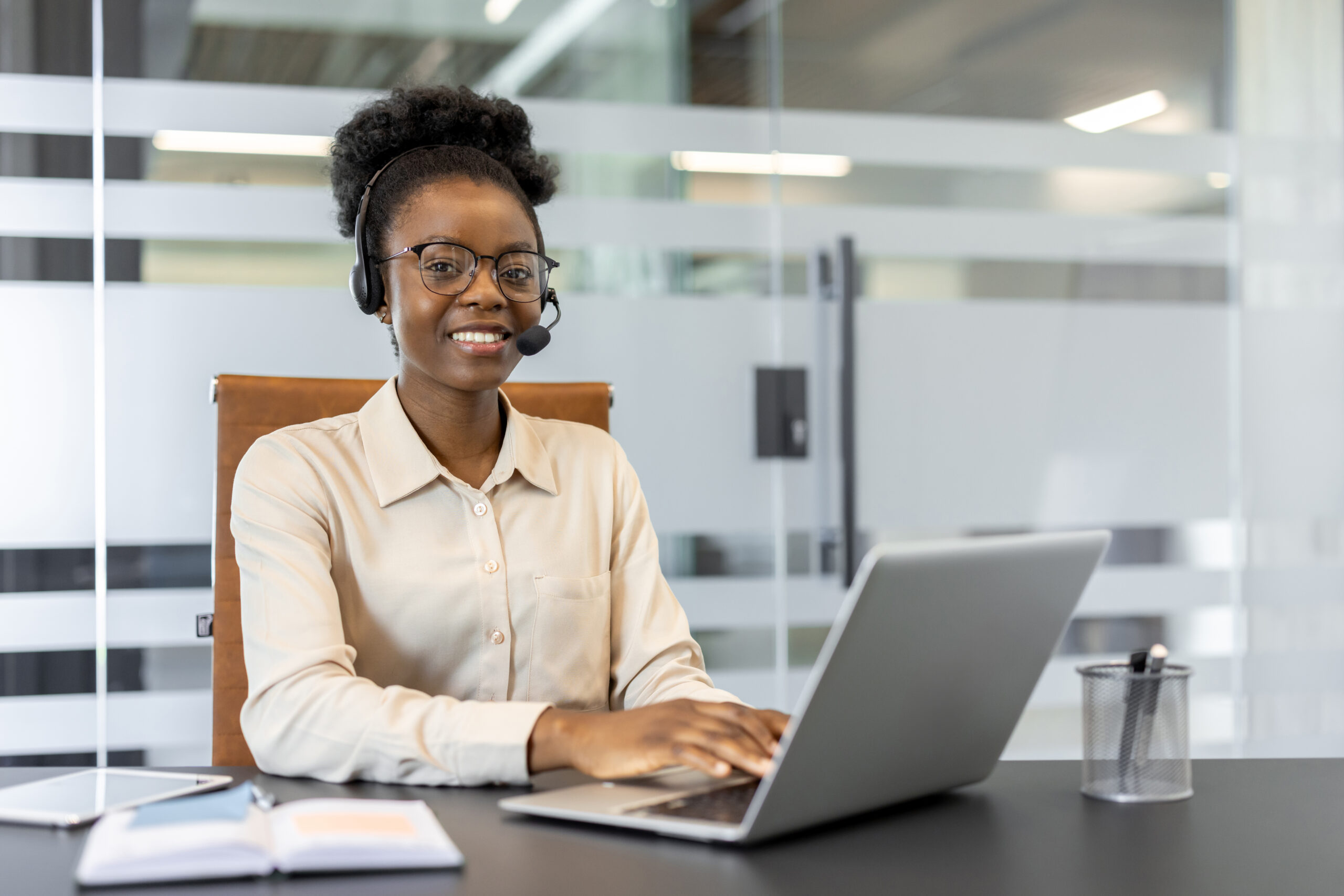 Confident African American businesswoman using laptop and headset for customer support. She is smiling, focused, and working efficiently in modern office setting.